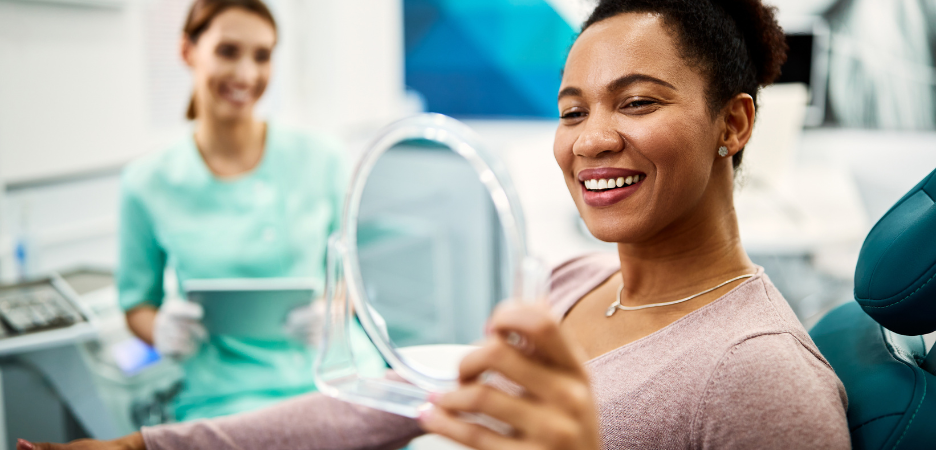 a black woman is in a dental chair, looking into a mirror and smiling showing off her white teeth. A female dentist is blurred in the background watching over her hard work.