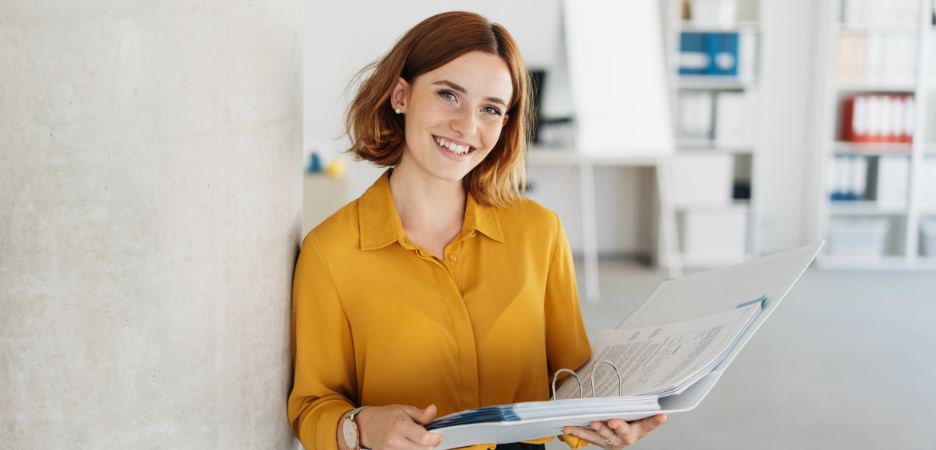 short haired, happy smiling ginger haired lady wearing an orange shirt holding a file binder