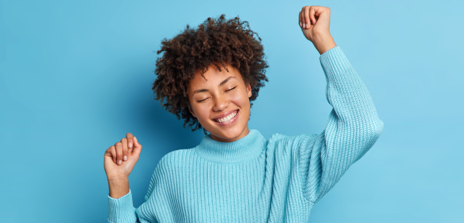 mixed race woman lifting hands in excitement with eyes closed wearing a blue top against a blue background
