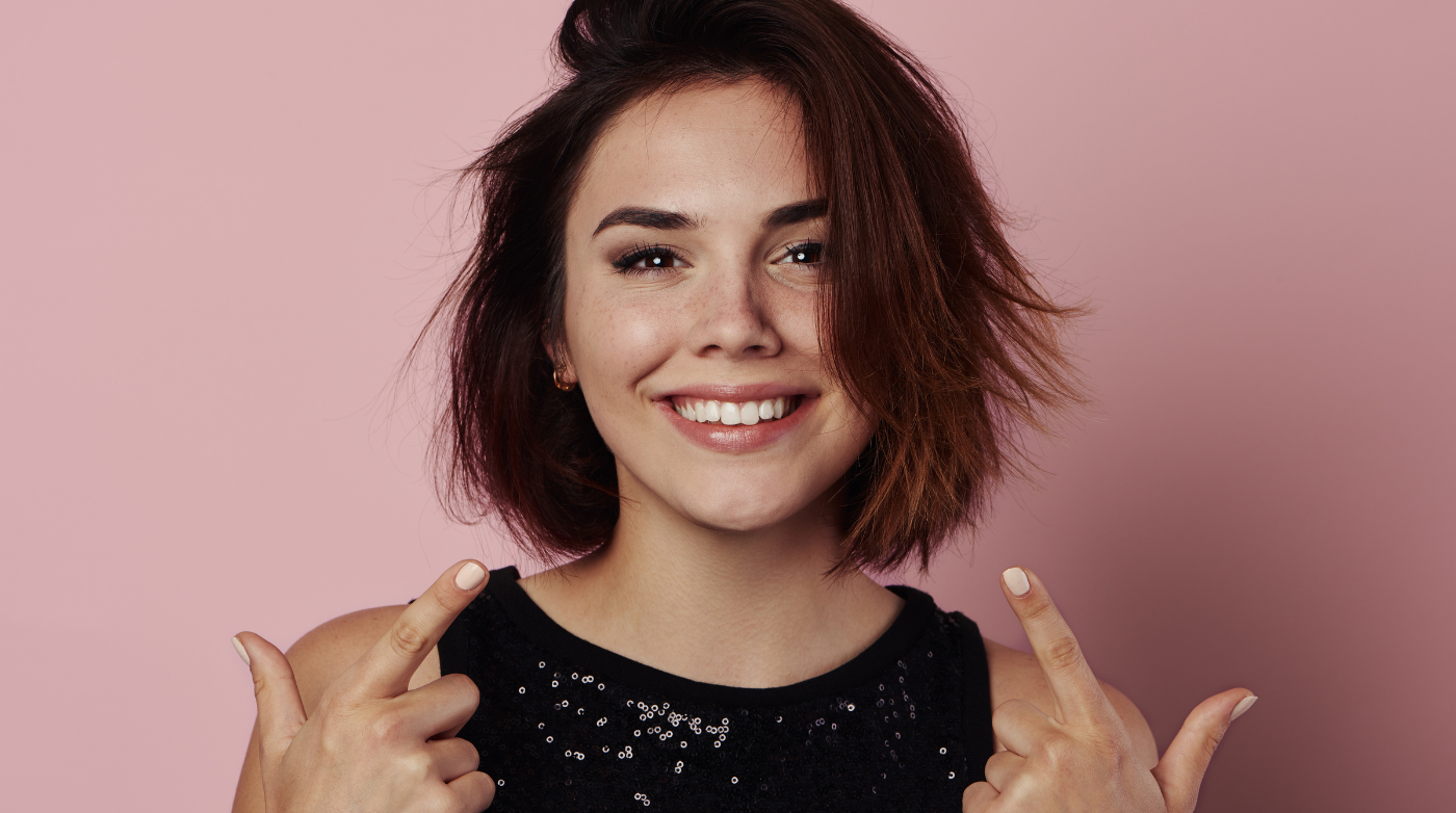 Woman smiling at camera and pointing at smile against a dark pink background